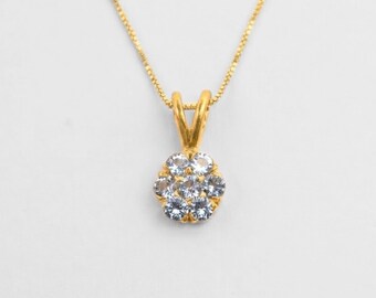 May include: A gold-colored necklace with a pendant featuring a cluster of small, clear gemstones. The pendant is flower-shaped, with the gemstones set closely together. The chain is delicate and also gold-colored, against a white background.