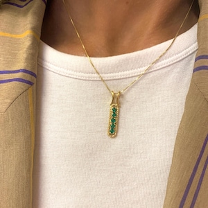 Op de afbeelding: Een gouden ketting met een hanger met een verticale staaf met zes groene edelstenen.
