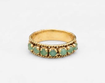 May include: A gold ring featuring a row of small, round, light green stones set between two twisted rope-style bands. The metal has a warm yellow tone, and the stones are evenly spaced along the circumference of the circular band design.
