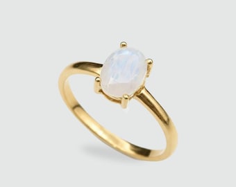 May include: A gold ring featuring an oval, iridescent gemstone. The ring's band and setting are a warm gold color, and the gemstone has a soft, milky appearance. The ring is a classic design, suitable for everyday wear or special occasions.