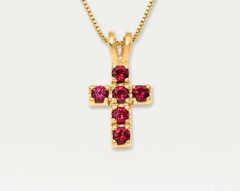 May include: A gold cross pendant necklace featuring five round, deep pink gemstones set in a vertical and horizontal arrangement. The pendant hangs from a delicate gold box chain, showcasing a polished metal finish and a classic, symmetrical design.