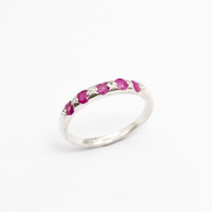 Puede incluir: Un anillo de banda color plateado que presenta cinco pequeñas piedras preciosas redondas de color rojo rosado vibrante dispuestas en fila en la parte superior. La banda de metal tiene un acabado liso y pulido, con engastes de metal.