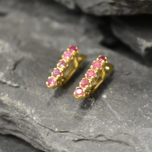 Puede incluir: Un par de pendientes de oro con una fila de piedras preciosas rosas. Los pendientes están diseñados en forma curva.