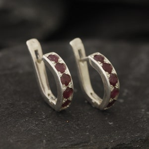 May include: A pair of silver hoop earrings with a row of red gemstones around the hoop.