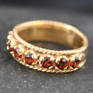 Pode incluir: Um anel de banda de ouro com um design trançado e uma fileira de pedras preciosas vermelhas.