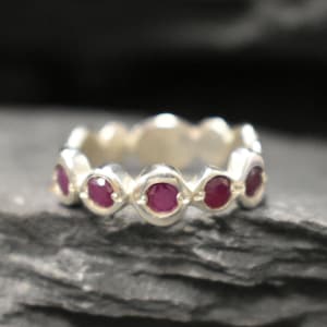 May include: A silver band ring with a row of six round red ruby gemstones.