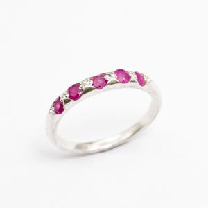May include: A silver ring featuring a line of oval-cut, vivid red gemstones. The gemstones are set closely together, creating a continuous line of colour. The ring is set against a plain white background.