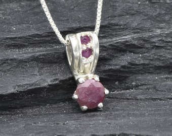 May include: A silver pendant necklace featuring a faceted, deep red ruby gemstone. The pendant has a decorative silver setting with two smaller rubies above the main stone. The necklace chain is a delicate silver box chain.