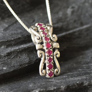 Könnte beinhalten: Eine silberne Halskette mit einem Anhänger in einem dekorativen Design mit einer Reihe roter Rubin-Steine.