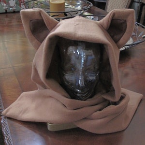 May include: A brown fleece hooded scarf with large, floppy ears. The scarf is folded and draped over a wooden surface.