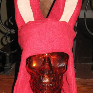 May include: A red fleece bunny hat with long, floppy ears. The ears are lined with a light brown fabric. The hat is sitting on a wooden surface.