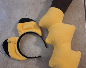 May include: A black headband with two yellow and black plush ears attached, alongside a separate yellow and brown plush lightning bolt-shaped tail. The items are made of a soft, fuzzy fabric and feature a bright yellow colour scheme with dark accents.