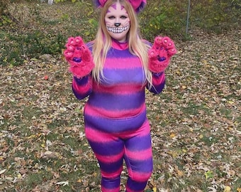 May include: A purple and pink striped Cheshire Cat costume. The costume includes a jumpsuit, gloves, and a headpiece with large ears. The face is painted with a wide grin. The costume is designed for Halloween or cosplay.