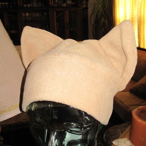 May include: A beige fleece hat featuring two prominent, upright, triangular cat-like ears on the top. The hat is constructed from a soft, textured fabric and is displayed on a dark, translucent mannequin head to show its shape and structure.