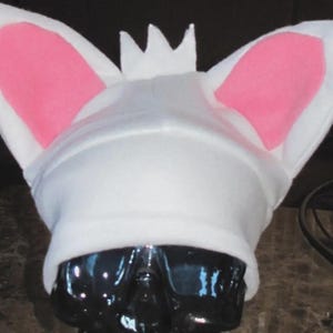 May include: A white beanie hat featuring two large, upright, pointed ears with pink inner sections and a small, three-pointed white crown centered between the ears. The hat is constructed from a soft, solid white fabric material.