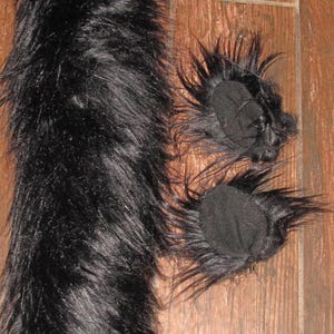May include: Black faux fur tail and two matching ear pieces. The tail is long and fluffy, and the ear pieces are round with a black fabric base and tufts of fur around the edges. The items are on a wooden surface.