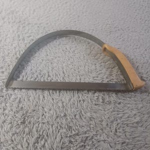 Puede incluir: Una sierra de calar con una hoja de metal y un mango de madera. La hoja de sierra es de color gris oscuro y tiene pequeños dientes. El mango es de color marrón claro.