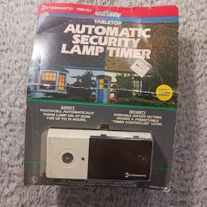 Op de afbeelding: Een vintage Intermatic automatische veiligheidslamp-timer voor op tafel in de originele verpakking. De verpakking is voornamelijk zwart, rood en blauw, met de tekst "Automatic Security Lamp Timer". De timer zelf is wit en zwart.
