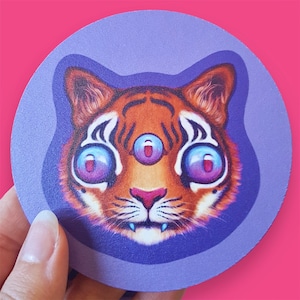 May include: A purple and white coaster featuring a cartoon illustration of a tiger with three eyes. The tiger has orange and black stripes and blue eyes.
