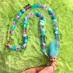 Puede incluir: Un collar de cuentas colorido con un colgante de muñeca troll vintage. El collar tiene cuentas azules, moradas y transparentes en forma de estrella, flor y oso. La muñeca troll tiene el pelo azul y una cara sonriente.