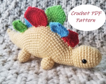 May include: A crocheted stegosaurus dinosaur in beige with red, green, and blue plates along its back. The dinosaur has a black eye and is lying on a gray, textured surface. Text on the image reads "Crochet PDF Pattern."
