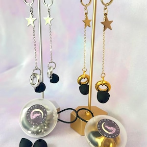 May include: Earrings with star charms and chain accents, displayed on silver and gold stands. The earrings are paired with black earbud tips and clear plastic containers with the text "Studio New Moon Magic".