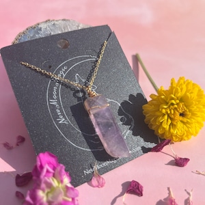 May include: A gold chain necklace with a purple amethyst crystal pendant. The pendant is wrapped in gold wire and has a raw, natural look. The necklace is displayed on a black card with the text "New Moon Magic" and a website address.