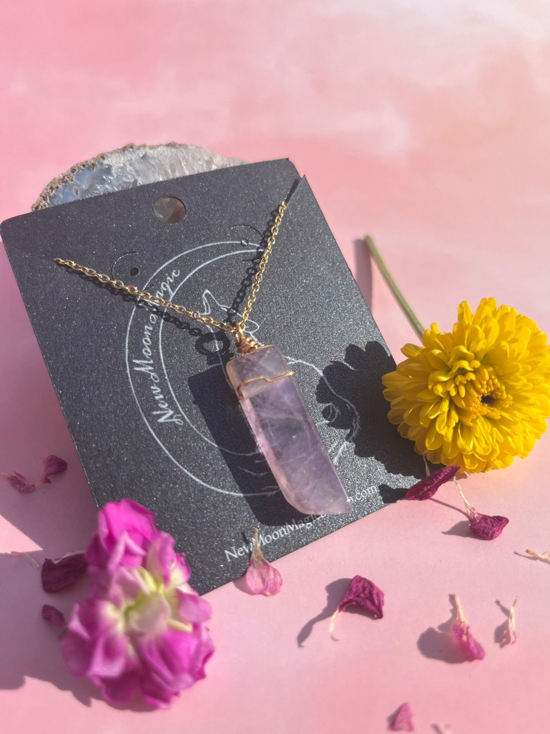 May include: A gold chain necklace with a purple amethyst crystal pendant. The pendant is wrapped in gold wire and has a raw, natural look. The necklace is displayed on a black card with the text "New Moon Magic" and a website address.