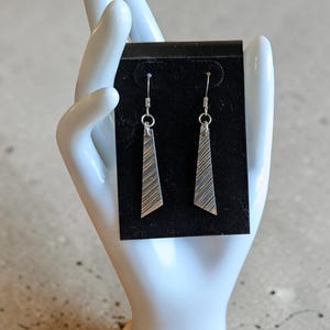 May include: A pair of silver-toned dangle earrings featuring a trapezoidal shape with a textured surface of diagonal etched lines. The earrings are suspended from silver-toned metal hooks and are displayed on a rectangular black card.