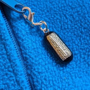 May include: A black and gold charm with a silver clasp attached to a blue zip pull. The charm is cylindrical with gold glitter embedded in the black glass. The background is a blue fleece fabric.