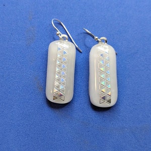 May include: A pair of silver earrings with white glass teardrop-shaped pendants. The pendants have a geometric pattern of iridescent triangles.