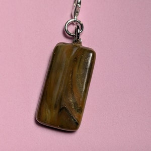 May include: A brown and gold rectangular charm with a silver clasp. The charm has a swirled pattern and a glittery finish.