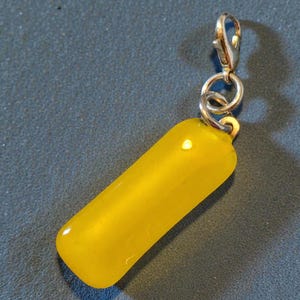 May include: A bright yellow, oblong charm with a silver-colored clasp. The charm is smooth and rounded, with a slight curve. The clasp is attached with a small silver ring. The charm is likely a pendant or accessory.