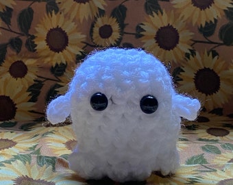May include: A small, white, crocheted ghost figure with a rounded body, two small outstretched arms, and two large, round, black plastic eyes. The texture of the yarn is visible throughout the surface of the figure, which has a soft, fuzzy appearance.