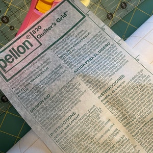 May include: A close-up shot of a quilting project in progress, featuring a pink rotary cutter and a ruler resting on a green cutting mat. The focus is on a sheet of Pellon Quilter's Grid with instructions in English and Spanish.