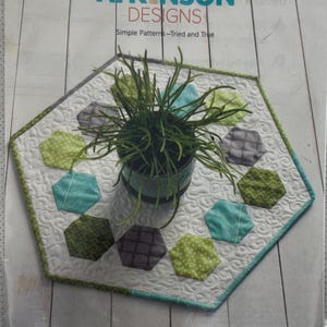 May include: A quilted hexagon table runner with a white background and colorful hexagon accents in shades of green, blue, and gray. The runner has a teal border and a small potted plant in the center. The text "MERRY-GO-ROUND" is visible.