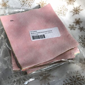 May include: A stack of pink, brown, and beige fabric squares in a clear plastic bag. The label on the bag reads "Kit Id: 5635 Quilter's Candy Flannel Soft Charms Charm Pack Fab Layers 14".