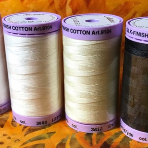 May include: Four spools of thread, two white, one cream, and one black. The spools are labeled "FINISH COTTON Art.9104" and "BLK-FINISH COTTON Art.9104".