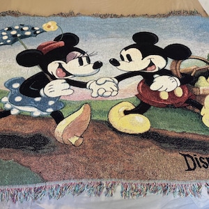 K&ouml;nnte beinhalten: Gewebte &Uuml;berwurfdecke mit Mickey Mouse und Minnie Mouse, die sich an den H&auml;nden halten. Die Decke hat unten ein Disneyland Resort-Logo. Die Decke hat einen Fransenrand und besteht aus einem weichen, gewebten Material.