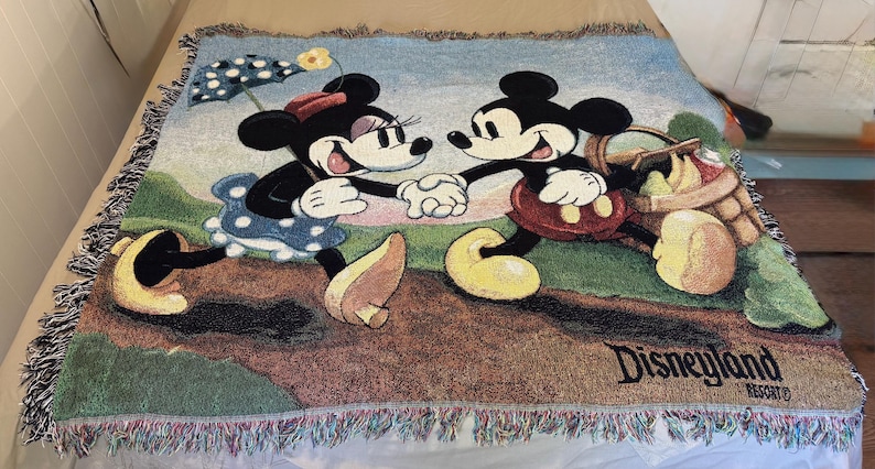K&ouml;nnte beinhalten: Gewebte &Uuml;berwurfdecke mit Mickey Mouse und Minnie Mouse, die sich an den H&auml;nden halten. Die Decke hat unten ein Disneyland Resort-Logo. Die Decke hat einen Fransenrand und besteht aus einem weichen, gewebten Material.