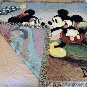 K&ouml;nnte beinhalten: Eine gewebte Disney-Wurfdecke mit Mickey und Minnie Mouse. Mickey tr&auml;gt einen Picknickkorb, und Minnie ist unter einem Polka-Dot-Schirm. Die Decke hat einen Fransenrand und das Wort "Disney" ist sichtbar.