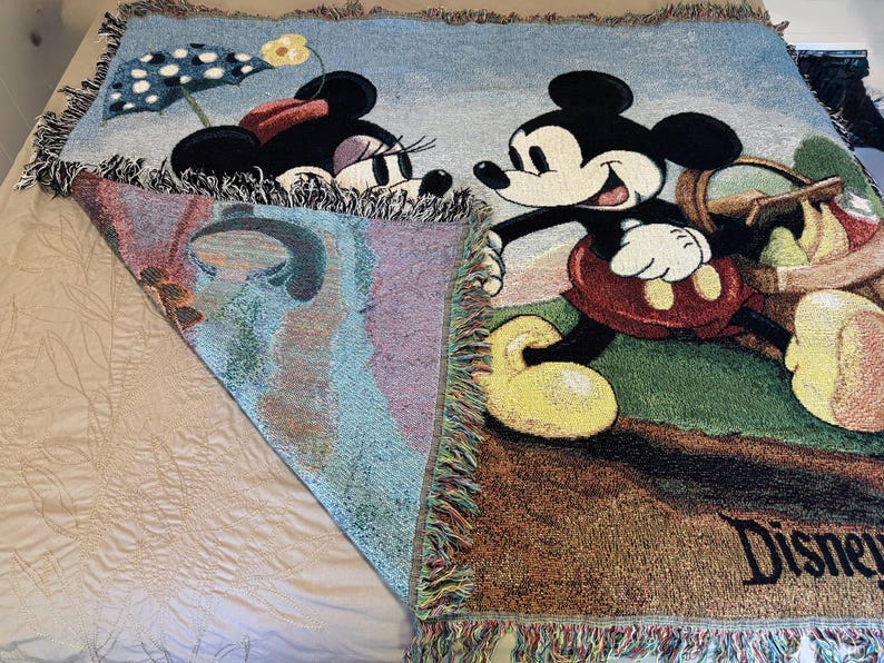 K&ouml;nnte beinhalten: Eine gewebte Disney-Wurfdecke mit Mickey und Minnie Mouse. Mickey tr&auml;gt einen Picknickkorb, und Minnie ist unter einem Polka-Dot-Schirm. Die Decke hat einen Fransenrand und das Wort "Disney" ist sichtbar.