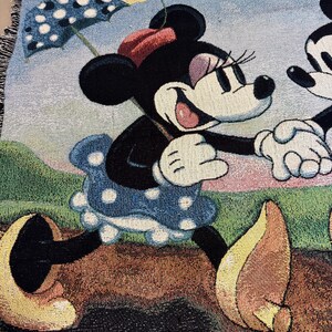K&ouml;nnte beinhalten: Gewebte Wurkdecke mit Minnie Mouse in einem blauen Polka-Dot-Kleid, die einen Polka-Dot-Regenschirm h&auml;lt. Die Decke hat einen hellblauen Himmelshintergrund mit einer gelben Blume und gr&uuml;nem Gras.