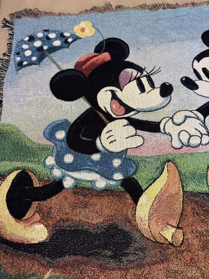 K&ouml;nnte beinhalten: Gewebte Wurkdecke mit Minnie Mouse in einem blauen Polka-Dot-Kleid, die einen Polka-Dot-Regenschirm h&auml;lt. Die Decke hat einen hellblauen Himmelshintergrund mit einer gelben Blume und gr&uuml;nem Gras.