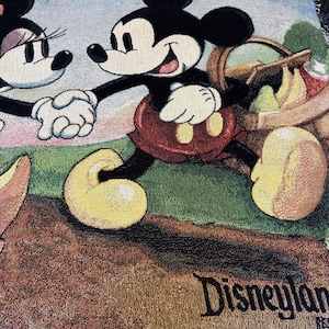 K&ouml;nnte beinhalten: Gewebte Tagesdecke mit Mickey Mouse und Minnie Mouse, die sich an den H&auml;nden halten, mit einem Picknickkorb. Die Decke hat ein Disneyland-Logo und einen Fransenrand. Die Farben sind Schwarz, Rot, Gelb, Gr&uuml;n und Blau.