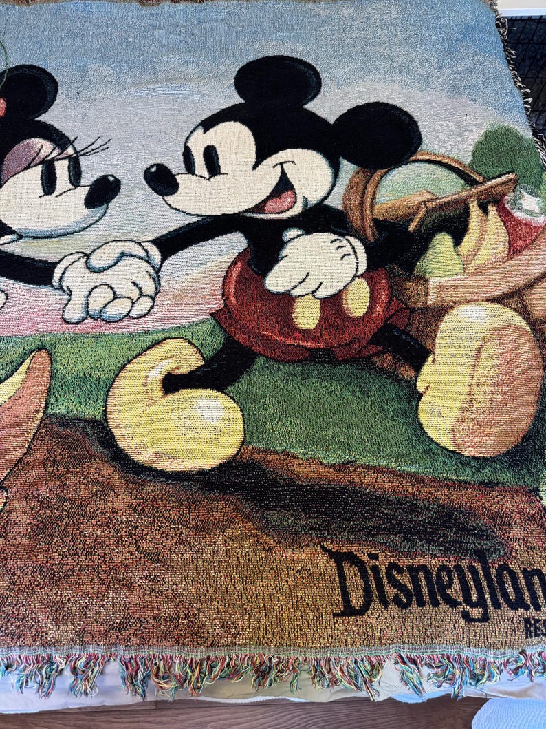 K&ouml;nnte beinhalten: Gewebte Tagesdecke mit Mickey Mouse und Minnie Mouse, die sich an den H&auml;nden halten, mit einem Picknickkorb. Die Decke hat ein Disneyland-Logo und einen Fransenrand. Die Farben sind Schwarz, Rot, Gelb, Gr&uuml;n und Blau.
