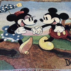 K&ouml;nnte beinhalten: Eine gewebte Decke mit Mickey Mouse und Minnie Mouse, die sich an den H&auml;nden halten. Minnie Mouse tr&auml;gt ein blaues Polka-Dot-Kleid und h&auml;lt einen Sonnenschirm. Mickey Mouse tr&auml;gt rote Shorts und tr&auml;gt einen Picknickkorb. Das Disney-Logo ist sichtbar.