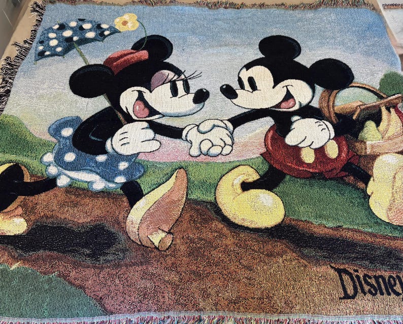 K&ouml;nnte beinhalten: Eine gewebte Decke mit Mickey Mouse und Minnie Mouse, die sich an den H&auml;nden halten. Minnie Mouse tr&auml;gt ein blaues Polka-Dot-Kleid und h&auml;lt einen Sonnenschirm. Mickey Mouse tr&auml;gt rote Shorts und tr&auml;gt einen Picknickkorb. Das Disney-Logo ist sichtbar.