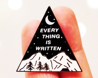 May include: A black and white enamel pin in a triangular shape. The pin features a mountain range, trees, a crescent moon, and the text "EVERY THING IS WRITTEN" in white. The pin is held between two fingers.