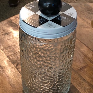 May include: A clear glass jar with a textured surface and a white lid. The lid has a black and white checkered pattern and a black knob on top.
